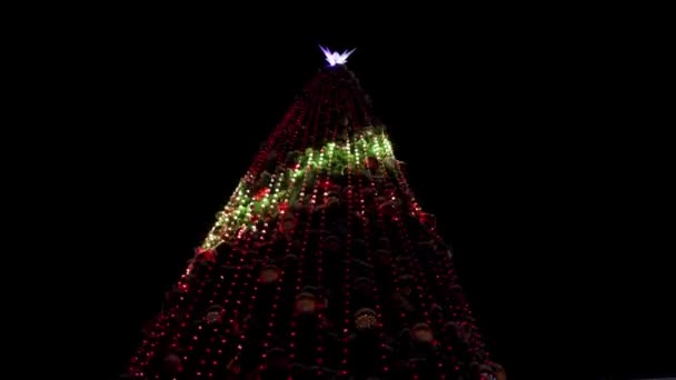 冬の雪の夜に街の広場に光輝くガーランドで飾られた大きなクリスマスツリー。カメラは通りの屋外の装飾でいっぱいの輝く新年の木の周りを飛んでいます。ライトショー. — ストック動画