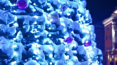 Karlı kış gecelerinde şehir meydanında parlak çelenkleri ve süslemeleri olan büyük bir Noel ağacı. Yakın plan. Dışarıdaki dekorasyonlarla dolu yeni yıl ağacının etrafında kameralar uçuşuyor. Işık Gösterisi