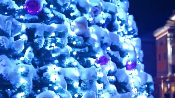 Grande árvore de Natal com guirlandas luminosas e decorações na praça da cidade na noite de inverno nevado. Close-up. A câmera voa ao redor da árvore de Ano Novo decorada brilhante cheia de decorações ao ar livre. Mostra de luz — Vídeo de Stock