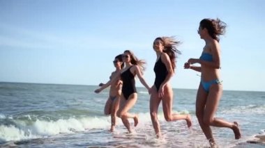 Bir grup güzel bayan arkadaş deniz kenarında mayo giymiş tropik kumsalda koşuyorlar. Bikinili ırklar arası kadınlar yaz günü açık havada bronzlaşır. Mayolu kızlar ağır çekimde koşar..