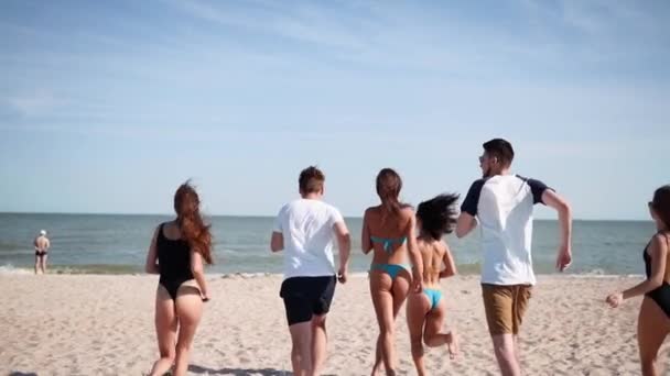 Grupo de amigos felices corriendo al agua de mar y saltando en la playa de arena. Gente alegre divirtiéndose en la playa en el día soleado de verano. Hombres y mujeres jóvenes van a nadar al océano. 120 fps cámara lenta . — Vídeo de stock