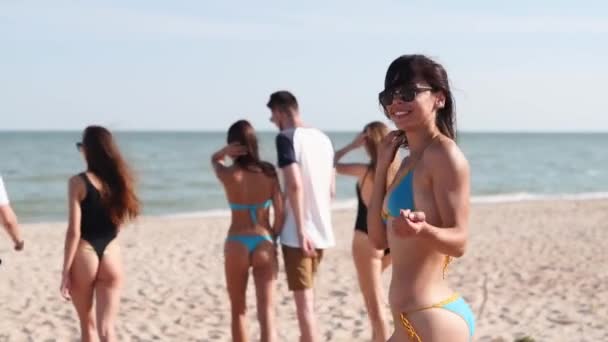 ビキニ姿の可愛い女性がカメラを向けると海で泳ぐ。ビーチでの水着の女の子のバックビューでは、オンにし、参加するために招待、従う。友達はゆっくりと海の岸に行く. — ストック動画