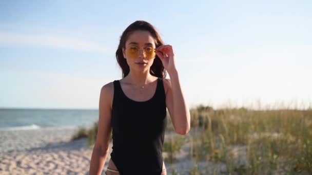 穿着泳衣和太阳镜在阳光灿烂的热带海滩上散步的年轻迷人女人。身穿比基尼的胖胖的漂亮姑娘慢吞吞地在沙滩上散步。太阳耀斑. — 图库视频影像