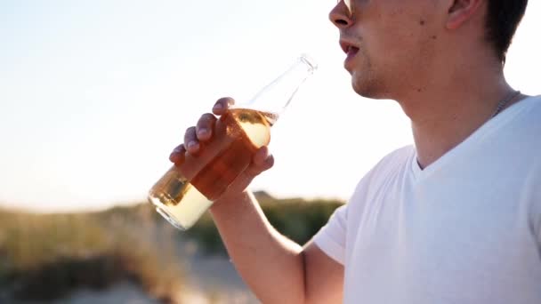Fiatal, jóképű fehér férfi, üveg sört iszik a parton, naplementekor, lassított felvételen. A hím kioltja a szomjat limonádéval a homokos tengerparton. Napkitörések, nyomkövető lövés.. — Stock videók