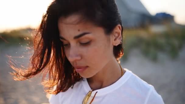 Retrato de uma jovem mulher olhando para a câmera em pé na praia com um mar no fundo. Menina atraente em t-shirt branca levanta os olhos. Vento sopra cabelos castanhos arejados brilhando em chamas do sol. — Vídeo de Stock