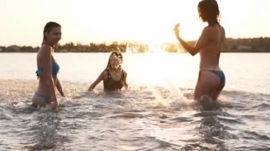 Bikinili mutlu kızlar gün batımında denize koşar ve birbirlerine su sıçratma oyunu oynarlar. Neşeli bayan arkadaşlar gölette su sıçratarak eğleniyorlar. Genç kadınlar yüzmeye gider, gölde yıkanırlar. Yavaş çekim.