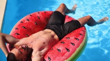 Şortlu adam yüzen karpuz su halkasının üzerinde dinleniyor. Arkadaşlar özel villa havuzunda şamandıraların üzerinde dinleniyorlar. Gençler güneşli bir günde lüks bir tatil köyündeki partide rahatlar. Yavaş çekim