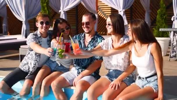 Група друзів весело проводять час на вечірці біля басейну клінкерні окуляри зі свіжими коктейлями, сидячи біля басейну в сонячний літній день. Люди тости п'ють напої на розкішній віллі на тропічних канікулах . — стокове відео