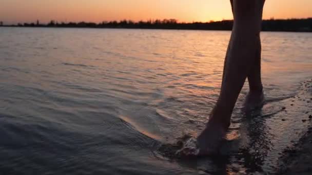 Manliga ben och fötter promenader längs sjön eller damm på sandstrand i skymningen. Män rör sig på land ensamma. Lugn vatten på bakgrunden. 120 fps slow motion. Killen går efter bad i bukten eller bäcken vid solnedgången. — Stockvideo