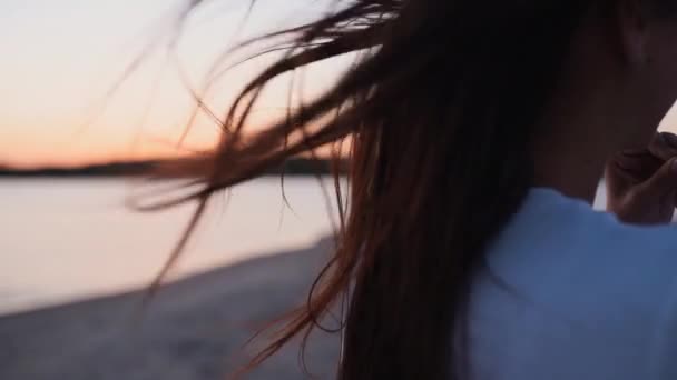 Chiudi indietro vista di marrone donna capelli tenero movimento in aria al tramonto estivo al rallentatore. Bruna o rossa donna in piedi su beachi al tramonto. Il vento soffia capelli ariosi che brillano all'alba. — Video Stock