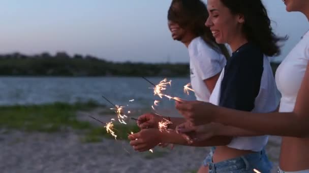 Amigas caminando, bailando, divirtiéndose en la fiesta nocturna en la playa con bengalas en las manos. Jóvenes adolescentes festejando en la playa con fuegos artificiales, luces de bengala. Las niñas en cámara lenta, steadycam disparo. — Vídeos de Stock