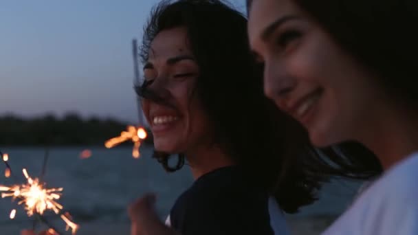 Amigos do sexo feminino andando, dançando, se divertindo na festa à noite à beira-mar com faíscas nas mãos. Jovens mulheres adolescentes festejando na praia com fogos de artifício, luzes de bengala. Meninas em câmera lenta, steadycam tiro. — Vídeo de Stock