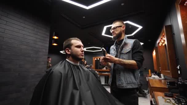 El peluquero moderno corta el cabello de los hombres barbudos con un cortador en la barbería. Para hombre peinado y corte de pelo en el salón. Aseo el cabello con trimmer. Peluquería haciendo corte de pelo en peluquería retro. Dolly en — Vídeo de stock