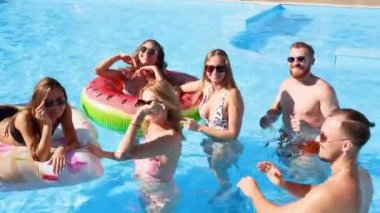 Arkadaşlar özel bir villada yüzme havuzunda parti yaparlar. Güneşli bir günde mayo giyen, dans eden, şamandıralarla takılan ve lüks bir tatil köyünde şişme yatak giyen mutlu gençler. İzleme görüntüsü.