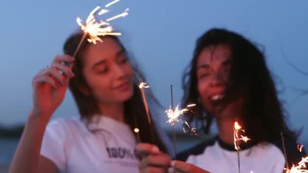 Des amies marchant, dansant, s'amusant à la soirée au bord de la mer avec des étincelles dans les mains. Jeunes adolescentes faisant la fête sur la plage avec feu d'artifice, lumières du bengale. Les filles au ralenti, steadycam shot. — Video