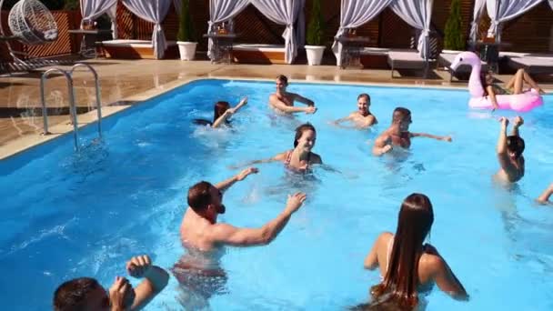 Friends have party in a private villa swimming pool. Happy young people in swimwear splashing water, dancing with floaties and inflatable mattress in luxury resort on sunny day. Slow motion.