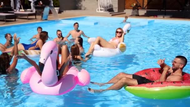 Przyjaciele relaksujący się w prywatnym basenie willi, leżą w słońcu na nadmuchiwanym flamingu, łabędziu, pływakach. Młodzi ludzie relaksują się na imprezie w luksusowym kurorcie w słoneczny dzień. Dziewczyny w bikini opalają się. Zwolniony ruch. — Wideo stockowe
