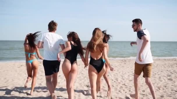 Groep van gelukkige vrienden rennend naar zeewater en springend op zandstrand. Vrolijke mensen die zich vermaken aan zee op de zonnige zomerdag. Jonge mannen en vrouwen gaan zwemmen in de oceaan. 120 fps slow motion. — Stockvideo
