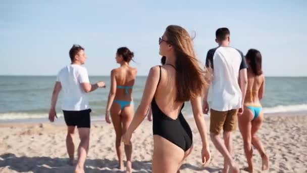 穿着比基尼的漂亮女人一边看着相机一边喊着要在海里游泳。穿着泳衣在海滩上散步的女孩的背影，转身邀请加入，跟随。朋友们慢吞吞地走到海滨去. — 图库视频影像