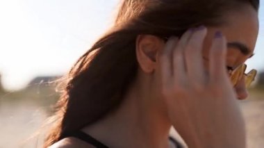Kahverengi kadın saçının yavaşça hareket edişini yakından izleyin. 120 fp. Kumsalda yürüyen güneş gözlüklü esmer kadın. Rüzgâr, günbatımında güneşte parlayan havadar saçlar savurur. Makro video.