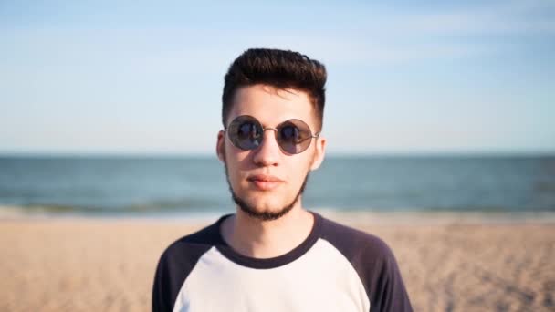 Πορτρέτο του νεαρού άνδρα που κοιτάζει και χαμογελά στην κάμερα που στέκεται στην παραλία με μια θάλασσα στο βάθος. Ευτυχισμένος τύπος με γυαλιά ηλίου κοιτάζοντας κάμερα χαλαρώνοντας στην ακτή του ωκεανού σε ηλιόλουστη μέρα σε εξωτερικούς χώρους. — Αρχείο Βίντεο