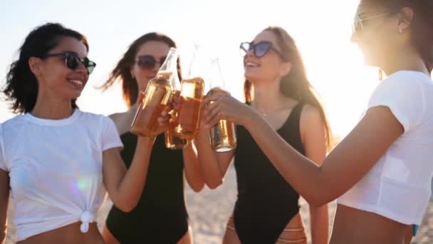 Hübsche Frauen im Bikini haben Spaß beim Anstoßen auf ein Getränk und entspannen am Sandstrand bei Sonnenuntergang. Fitte Mädchen in Badeanzügen, Sonnenbrillen, die mit Bierflaschen am Ufer hängen. Junge Frauen trinken Limonade. — Stockvideo