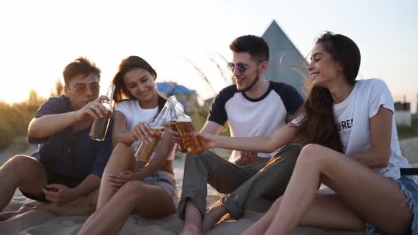 Group of friends have fun toasting, drinking beer, relaxing on sea beach at sunset in slow motion. Young men, women enjoy beverage sitting on a sand on warm summer evening party. People with lemonade. — Stock Video