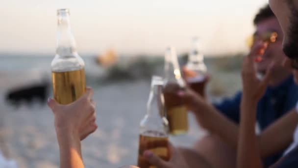 Skupina přátel se baví tousty, pití piva, relaxace na mořské pláži při západu slunce ve zpomaleném filmu. Mladí muži, ženy mají rády nápoje posazené na písku na teplém letním večírku. Lidé s limonádou. — Stock video