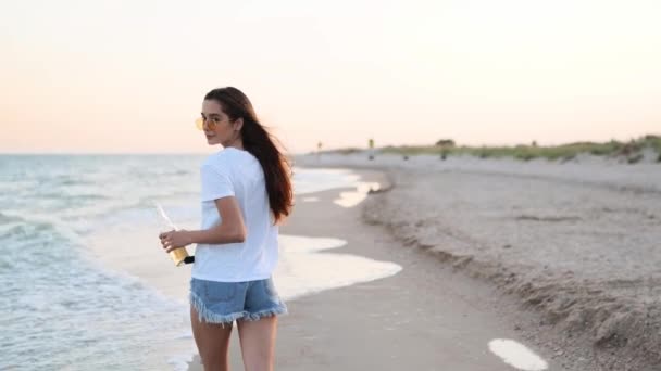 Vacker kvinna promenader längs havet på sandstrand har kul med öl flaska. Attraktivt utrustad flicka höjer händerna, njuter av fredom och sommarsemester nära havet stranden, dricka dryck. Långsamma rörelser. — Stockvideo