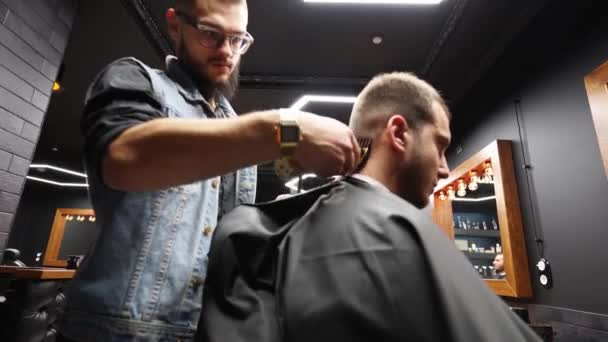Trendy barber cuts bearded mans hair with a clipper in barbershop. Mens hairstyling and hair cutting in salon. Grooming the hair with trimmer. Hairdresser doing haircut in retro hair salon. Dolly — Stock Video