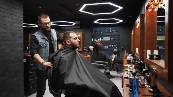 Hairdresser in glasses finishes a haircut and taking off salon cape from masculine client in barbershop. Barber turns chair and removes cloak from customer. Master and client look at camera. Dolly in. — Stock Video