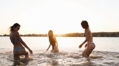 Bikinili mutlu kızlar gün batımında denize koşar ve birbirlerine su sıçratma oyunu oynarlar. Neşeli bayan arkadaşlar gölette su sıçratarak eğleniyorlar. Genç kadınlar yüzmeye gider, gölde yıkanırlar. Yavaş çekim.