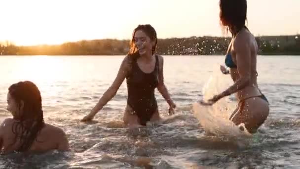 Glada våta tjejer i bikini springer ut i havet och leker stänk vatten till varandra vid solnedgången. Glada kvinnliga vänner har kul att göra stänk i dammen. Unga kvinnor badar, badar i sjön. Långsamma rörelser. — Stockvideo