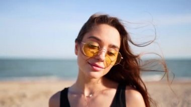 Sahilde duran arka planda deniz olan genç bir kadının portresi. Siyah mayo ve güneş gözlüğü takan çekici bir kız gözlerini açıyor. Rüzgar esiyor. Kahverengi saçlar güneşte parlıyor..