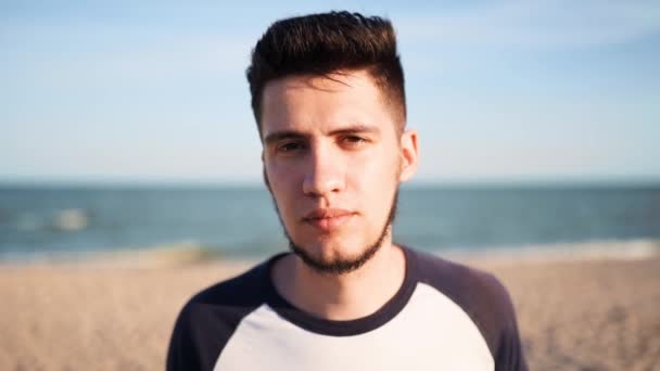 Close portrait of a young man with tired sad eyes looking at the camera standing on a beach with sea on background. Thoughtful wistful guy stares at viewer. Feelings and depression concept. — Stock Video