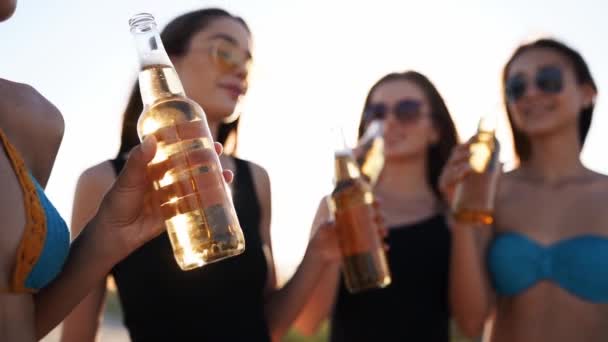 Hübsche Frauen im Bikini haben Spaß beim Anstoßen auf ein Getränk und entspannen am Sandstrand bei Sonnenuntergang. Fitte Mädchen in Badeanzügen, Sonnenbrillen, die mit Bierflaschen am Ufer hängen. Junge Frauen trinken Limonade. — Stockvideo