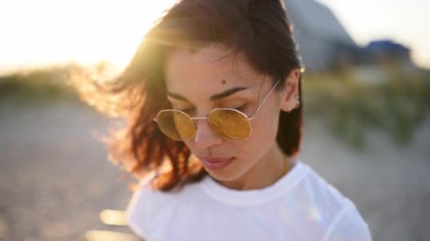 Ritratto di giovane donna che guarda la macchina fotografica in piedi sulla spiaggia con un mare sullo sfondo. Attraente ragazza in t-shirt bianca e occhiali da sole alza gli occhi. Il vento soffia ariosi capelli castani che brillano nei razzi del sole. — Video Stock