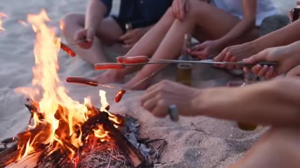 Közelről látjuk, ahogy a barátok kolbászt sütnek a tábortűz körül, sört isznak, gitároznak a homokos parton. Fiatal csoport férfiak és nők ital énekel együtt gitározni közel tábortűz alkonyatkor. — Stock videók