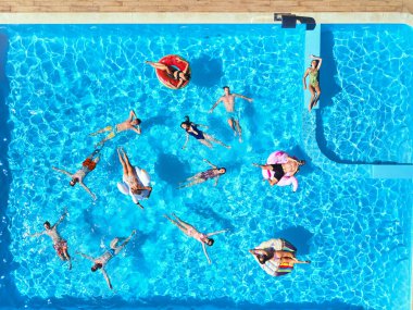 Havuzda, şişme flamingo, kuğu ve şiltelerle parti veren arkadaşlar. Mutlu gençler güneşli bir günde lüks bir tatil köyünde dinleniyorlar. Yukarıdan bak. Bikinili kızlar güneşte güneşleniyor..