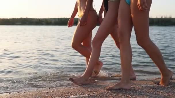 Slim piernas y pies femeninos caminando a lo largo del lago o estanque en la playa de arena. Las mujeres bonitas se mueven en la orilla. Agua calmada en el fondo. 120 fps en cámara lenta. Chicas colgando después de bañarse en la bahía o arroyo en la puesta del sol . — Vídeo de stock