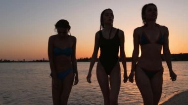 Gün batımında denizde banyo yaptıktan sonra bikinili ince ve güzel kızlar sudan çıkıyordu. Mayolu kadınlar göletten tropik plaja giderler. Gündoğumunda alacakaranlıkta bayan arkadaşlar. Yavaş çekim.