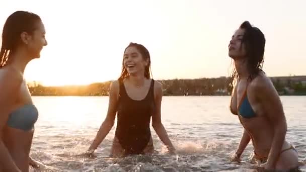 Glada våta tjejer i bikini springer ut i havet och leker stänk vatten till varandra vid solnedgången. Glada kvinnliga vänner har kul att göra stänk i dammen. Unga kvinnor badar, badar i sjön. Långsamma rörelser. — Stockvideo