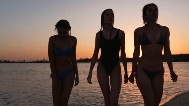 Fit slanke mooie meisjes silhouetten in bikini lopen uit het water na het baden in zee bij zonsondergang. Vrouwen in zwempakken gaan van vijver naar tropisch strand. Vrouwelijke vrienden bij zonsopgang. Langzame beweging. — Stockvideo