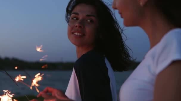 Amigas caminando, bailando, divirtiéndose en la fiesta nocturna en la playa con bengalas en las manos. Jóvenes adolescentes festejando en la playa con fuegos artificiales, luces de bengala. Las niñas en cámara lenta, steadycam disparo. — Vídeos de Stock