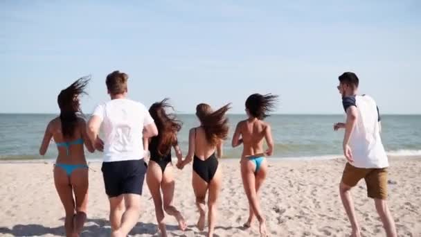 Groep van gelukkige vrienden rennend naar zeewater en springend op zandstrand. Vrolijke mensen die zich vermaken aan zee op de zonnige zomerdag. Jonge mannen en vrouwen gaan zwemmen in de oceaan. 120 fps slow motion. — Stockvideo