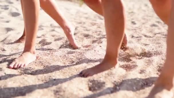 Vista cercana de las mujeres bronceadas piernas y pies caminando en la playa de arena hasta el océano en el día soleado. Chicas hermosas delgadas van por el mar en la arena blanca y transparente. Amigos de vacaciones. Ángulo de cámara bajo, cámara lenta. — Vídeos de Stock