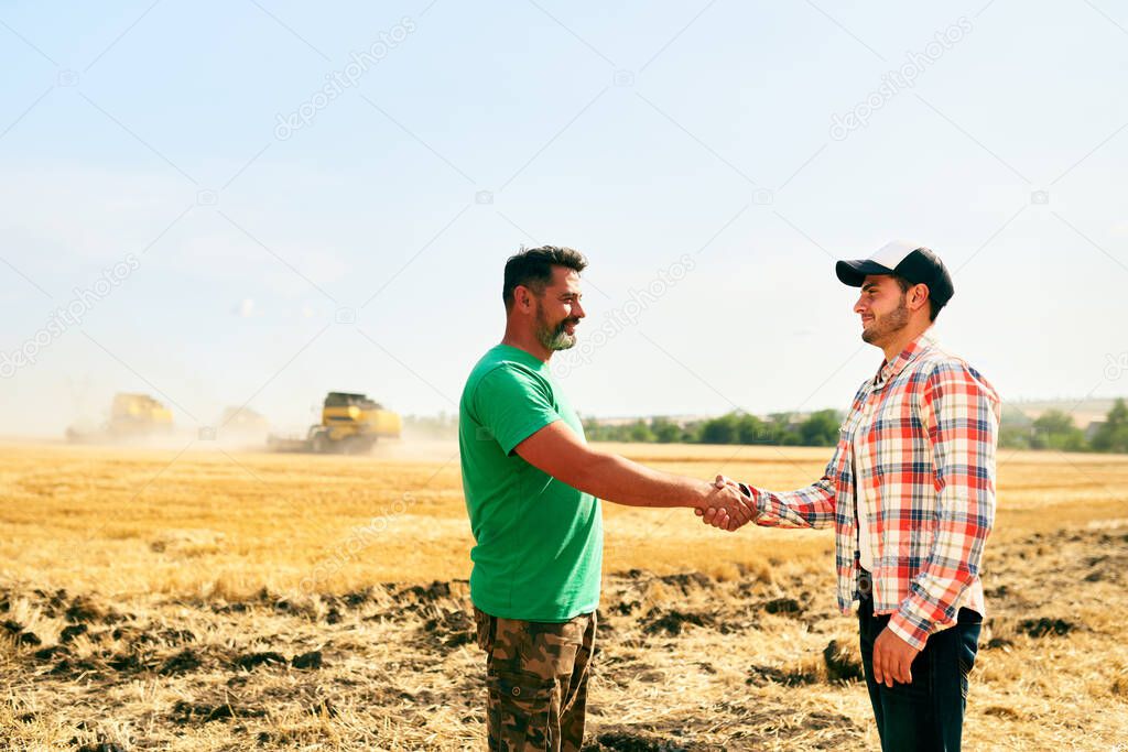 Farmer and agronomist shaking hands in wheat field after agreement. Agriculture business contract concept. Corporate farmer and landlord rancher negotiate with handshake. Combine harvesters harvest.