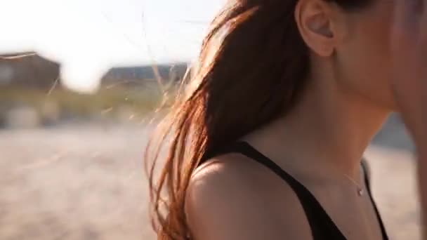 Закройте вид назад на нежное движение волос коричневой женщины в воздухе в замедленном режиме 120 кадров в секунду. Брюнетка в солнечных очках гуляет по песчаному пляжу. Ветер дует воздушные волосы, сияющие на солнце на закате. Макровидео. — стоковое видео