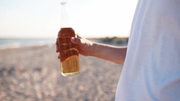 Jovem caucasiano bonito bebendo cerveja de garrafa de vidro na praia durante o pôr do sol em câmera lenta. Masculino sacia sede com bebida de limonada na costa do mar arenoso. Sinalizadores de sol, tiro de rastreamento. — Vídeo de Stock