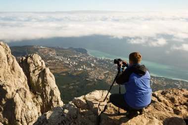 Bulutların, şehrin ve denizin üstündeki yüksek bir dağın tepesinde tripod ve fotoğraf makinesiyle oturan bir adam. Profesyonel fotoğrafçı kayalık zirvede DSR ayarlarını ayarlıyor.
